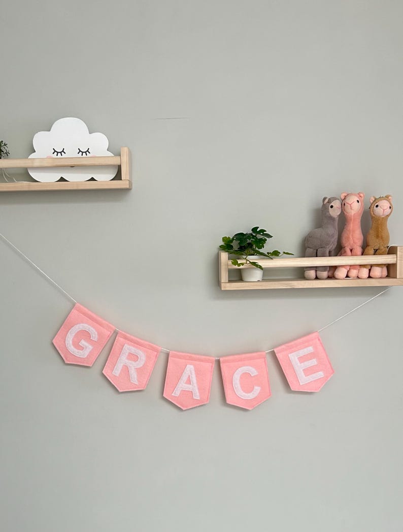 Puede incluir: Una pancarta de fieltro rosa con la palabra "GRACE" cuelga debajo de dos estantes de madera. El estante superior contiene una decoraci&oacute;n blanca en forma de nube con los ojos cerrados. El estante inferior muestra llamas de peluche.