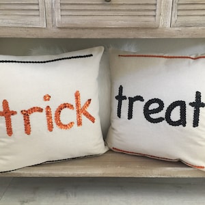 Puede incluir: Dos almohadas decorativas con las palabras "trick" y "treat" escritas en lentejuelas naranjas y negras. Las almohadas están en un estante de madera.