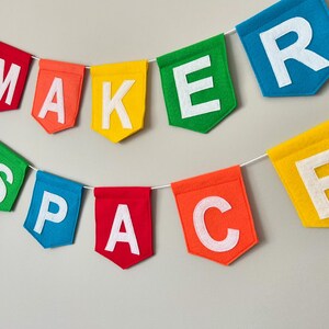 Puede incluir: Una colorida guirnalda de fieltro que deletrea "MAKER SPACE" con letras blancas. Las banderolas son rojas, naranjas, amarillas, verdes, azules y naranjas. La guirnalda est&aacute; ensartada en una cuerda blanca, perfecta para una sala de manualidades.