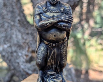 May include: A bronze-colored candle shaped like a horned demon with crossed arms, a cape with skull accents, and a belt. The base of the candle reads "MAMMON". The candle has a white wick.