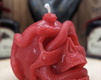 May include: A red skull candle with a snake wrapped around it. The candle has a white wick and is made of wax.