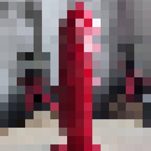 May include: A tall, red, abstract sculpture with a rounded base and a vertical, textured body. The sculpture is the focal point, set against a blurred background of neutral tones. The artwork appears to be made of a solid material.