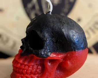 May include: A black and red skull-shaped candle with a white wick. The top half of the skull is black, and the bottom half is red.