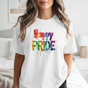 May include: A white short-sleeved t-shirt featuring the text Happy PRIDE in a multicolored watercolor style. The letters display a vibrant spectrum of red, orange, yellow, green, blue, and purple with paint drip effects.