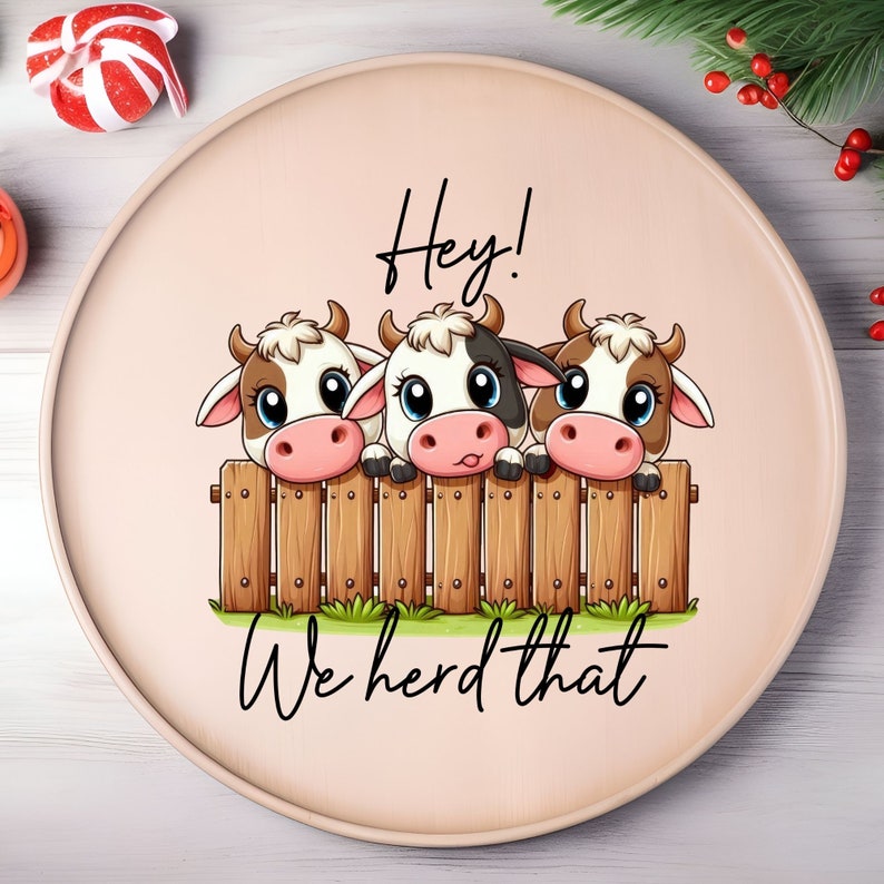May include: A round wooden platter with a pink background and the text "Hey! We herd that" in black. Three cartoon cows with brown and white spots are peeking over a wooden fence.