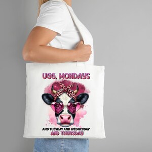 Op de afbeelding: Witte tote bag met een roze en zwarte koe die hartvormige zonnebrillen en een roze bandana draagt. De tekst "UGG, MONDAYS AND TUESDAY AND WEDNESDAY AND THURSDAY" staat op de tas gedrukt.