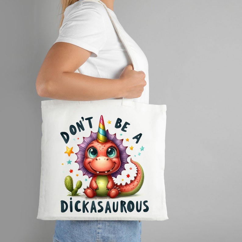 Puede incluir: Bolsa de tela blanca con un dinosaurio de dibujos animados colorido con un cuerno de unicornio. El texto en la bolsa dice "Don't be a Dickasaurus".