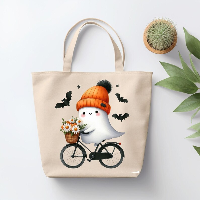 May include: A beige tote bag with a cute ghost wearing an orange and black hat riding a bicycle with a basket of white flowers. The ghost has a happy expression and there are black bats flying around it.