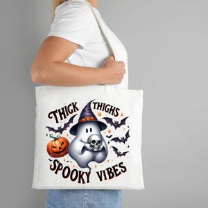 May include: White tote bag with a ghost wearing a witch hat and holding a skull. The bag has the text "Thick Thighs Spooky Vibes" printed on it.