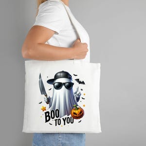 May include: White tote bag with a cartoon ghost wearing sunglasses and a baseball cap. The ghost is holding a knife and a pumpkin. The text "BOO TO YOU" is printed on the bag.