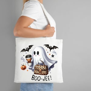 May include: White tote bag with a cartoon ghost holding a coffee cup and a leopard print purse. The ghost has big eyes and a cute expression. The bag also has bats and stars printed on it. The text "BOO-JEE!" is printed on the bottom of the bag.