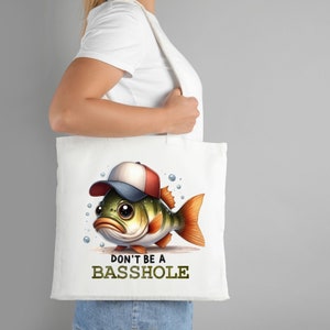 Puede incluir: Bolsa de tela blanca con un bajo de dibujos animados que lleva una gorra de b&eacute;isbol roja y blanca. El texto "DON'T BE A BASSHOLE" est&aacute; impreso debajo del pez.