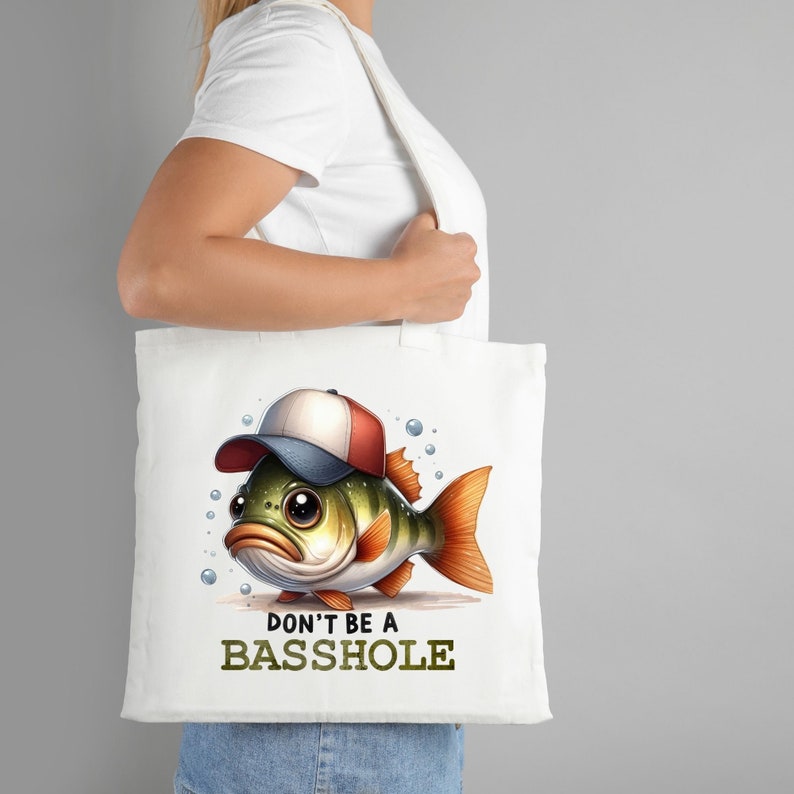 Puede incluir: Bolsa de tela blanca con un bajo de dibujos animados que lleva una gorra de b&eacute;isbol roja y blanca. El texto "DON'T BE A BASSHOLE" est&aacute; impreso debajo del pez.
