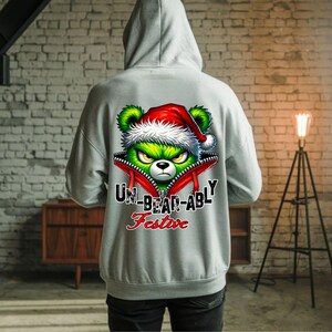 May include: A light gray hooded sweatshirt featuring a graphic on the back of a green bear wearing a red Santa hat, peeking out from a red zippered opening. The text reads UN-BEAR-ABLY Festive in black and red lettering.