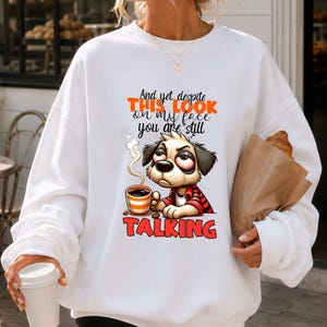 May include: A white long-sleeve sweatshirt featuring a graphic of a tired-looking dog holding a steaming coffee mug. The text reads: And yet despite THIS LOOK on my face you are still TALKING. The dog wears a red and black plaid shirt.