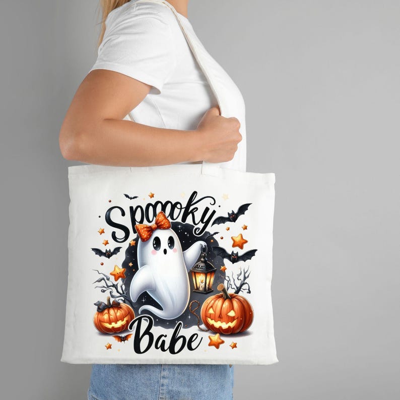 May include: White tote bag with a spooky Halloween design. The design features a ghost with a pink bow, a lantern, and pumpkins. The text "Spooky Babe" is printed on the bag.