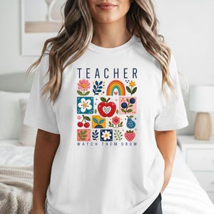 May include: A white t-shirt featuring the text TEACHER WATCH THEM GROW in dark blue letters. The graphic includes a grid of colorful illustrations including a rainbow, an apple, flowers, cherries, blueberries, and a strawberry.