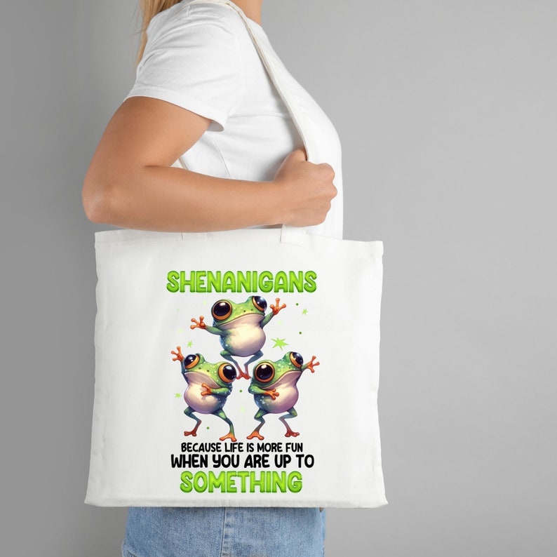 Op de afbeelding: Witte tote bag met een groen kikkerontwerp. De tekst op de tas luidt "Shenanigans because life is more fun when you are up to something."