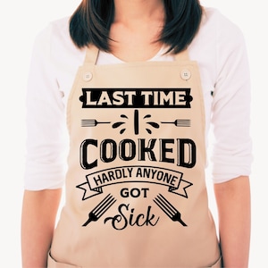 Może przedstawiać: Beżowy fartuch z czarnym tekstem, kt&oacute;ry brzmi "Last time I cooked hardly anyone got sick". Tekst jest otoczony czarną wstążką i dwoma czarnymi widelcami.
