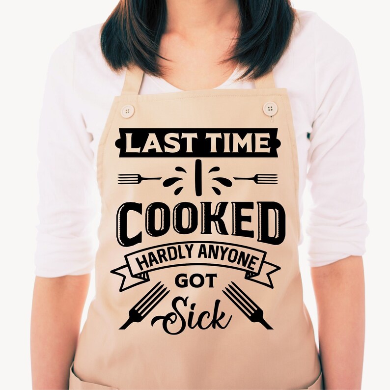 Może przedstawiać: Beżowy fartuch z czarnym tekstem, kt&oacute;ry brzmi "Last time I cooked hardly anyone got sick". Tekst jest otoczony czarną wstążką i dwoma czarnymi widelcami.