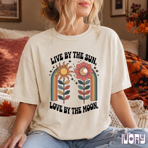 May include: An ivory short-sleeve t-shirt featuring a graphic print with the text "LIVE BY THE SUN, LOVE BY THE MOON," a yellow sun, a pink flower with a crescent moon center, and colorful rainbow arches with green leaves.