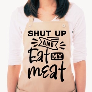 Może przedstawiać: Beżowy fartuch z czarnym napisem "Shut Up And Eat My Meat".