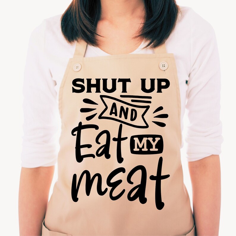 Może przedstawiać: Beżowy fartuch z czarnym napisem "Shut Up And Eat My Meat".