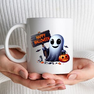K&ouml;nnte beinhalten: Wei&szlig;e Keramik-Kaffeetasse mit einem Cartoon-Geist, der ein Schild mit der Aufschrift "Happy Halloween" und einen K&uuml;rbis h&auml;lt. Der Geist l&auml;chelt und hat gro&szlig;e Augen.