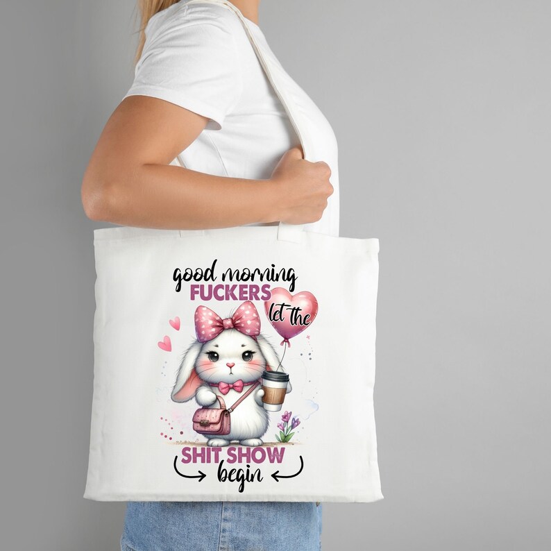 Op de afbeelding: Witte tote bag met een roze konijn dat een kopje koffie en een roze hartvormige ballon vasthoudt met de tekst "let the shit show begin". Het konijn draagt een roze strik en een roze handtas. De tekst op de tas is "good morning fuckers let the shit show begin".