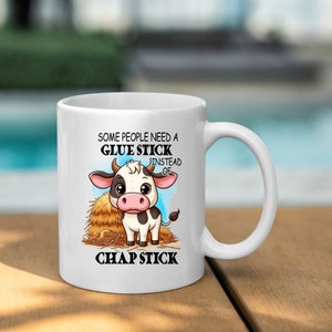 May include: White ceramic mug with a cartoon cow and the text "Some people need a glue stick instead of chap stick".