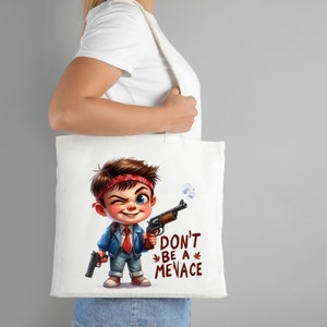 Puede incluir: Bolsa de tela blanca con una ilustraci&oacute;n de dibujos animados de un ni&ntilde;o que lleva un pa&ntilde;uelo rojo, una chaqueta azul y una corbata roja. Est&aacute; sosteniendo un arma en cada mano y tiene una expresi&oacute;n traviesa en su rostro. El texto "Don't be a menace" est&aacute; impreso debajo de la ilustraci&oacute;n.