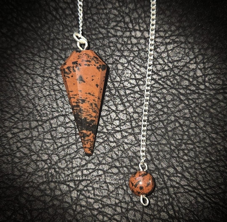 May include: A pendulum with a faceted, reddish-brown stone, speckled with black, suspended from a silver chain. A smaller, round stone of the same color and pattern hangs at the chain's end. The pendulum is designed for divination and spiritual practices.