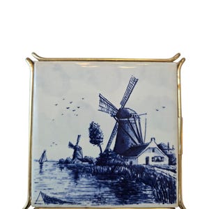 May include: A square ceramic tile featuring a blue and white landscape illustration of two windmills, a house, and a sailboat on water. The tile is encased in a thin, polished gold-toned metal frame with curved corners that extend outward.