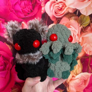 Puede incluir: Dos juguetes de peluche de crochet, uno negro con pelo marrón y ojos rojos, el otro verde con ojos rojos. Los juguetes se sostienen en una mano sobre un fondo de rosas rosas.