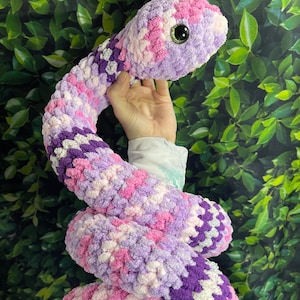 Puede incluir: Un juguete de peluche de serpiente tejido a crochet en tonos de morado, rosa y blanco. La serpiente tiene un ojo grande, negro con un iris verde.