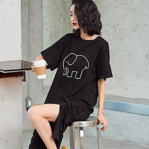 Peut inclure: Une robe t-shirt noire avec un contour blanc d'un éléphant sur le devant. La robe a une fente sur le côté et le mannequin tient une tasse de café.