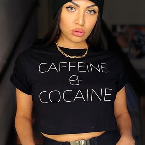 May include: A woman wearing a black crop top with the text "CAFFEINE or COCAINE" in white. She is also wearing a black beanie with a white logo, a gold chain necklace, a gold belt, and a gold watch.