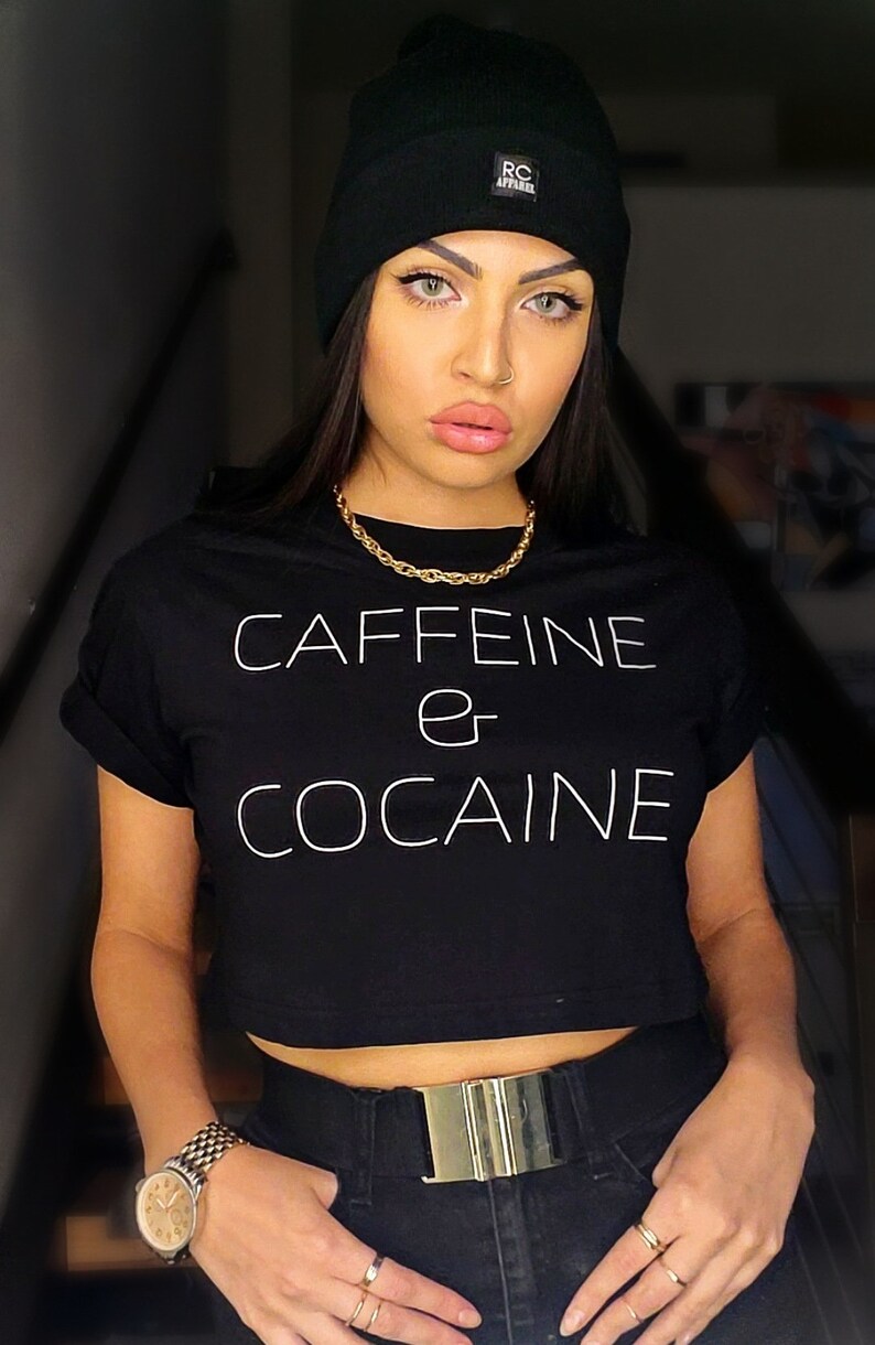 May include: A woman wearing a black crop top with the text "CAFFEINE or COCAINE" in white. She is also wearing a black beanie with a white logo, a gold chain necklace, a gold belt, and a gold watch.