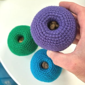 Puede incluir: Tres juguetes para perros de ganchillo de colores brillantes en tonos morado, turquesa y verde. Cada juguete en forma de rosquilla tiene una abertura central llena de pequeñas golosinas marrones. Estos juguetes están diseñados para el enriquecimiento y el juego de mascotas.