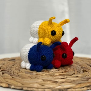 May include: Three crocheted stuffed toys shaped like bugs. The top bug is yellow and white, the middle bug is blue and white, and the bottom bug is red and white.