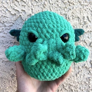 May include: A handmade, teal-colored crochet Cthulhu plushie with large black eyes and small, dark green wings. The plushie has a textured, rounded body and tentacles. The toy is held in a hand.