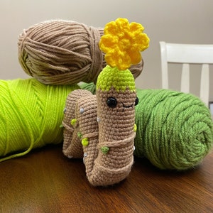 May include: A crocheted amigurumi cactus toy with a yellow flower on top. The cactus is brown with green accents and has a smiling face. The toy is surrounded by balls of yarn in brown, green, and yellow.