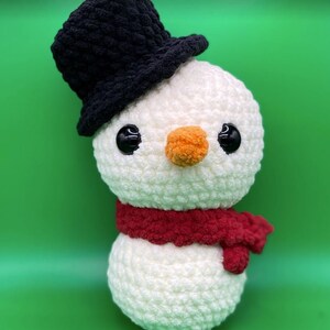 May include: A white crocheted snowman with a black top hat and a red scarf. The snowman has black eyes and an orange nose.