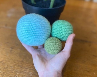 May include: Three crocheted balls, one light blue and two green, are held in a hand. The balls are different sizes.