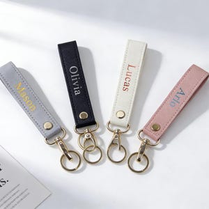 May include: Four personalised keychains in grey, black, white, and pink. Each keychain has a name embroidered in a contrasting colour: Mason, Olivia, Lucas, and Arlo. The keychains have gold-coloured hardware.