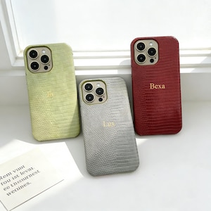 May include: Three phone cases in shades of green, gray, and red. Each case has a textured, reptile-like pattern and a gold-colored camera ring. The red case has the name "Bexa" printed on it, and the other two have initials.