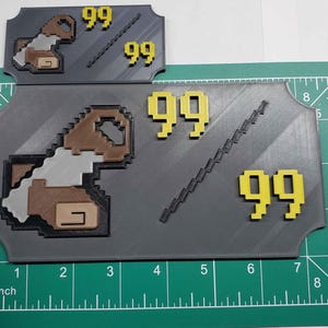 Op de afbeelding: Een grijs 3D-geprint bord met een gepixelde afbeelding van een bruin en wit wezen en de nummers 99 in het geel gedrukt. Het bord ligt op een groene snijmat met inchmarkeringen.