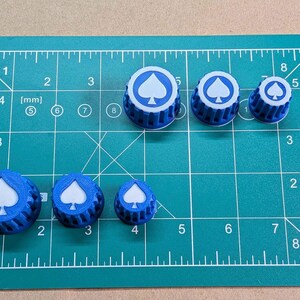 May include: Six blue circular knobs with a white spade symbol on the top surface, arranged in two rows on a green cutting mat. The knobs feature a ribbed side texture and vary in size, with the largest measuring approximately 1 inch in diameter.