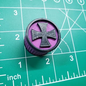 Op de afbeelding: Een paars-zwarte plastic flesdop met een zwart kruisontwerp. De dop ligt op een groene snijmat met een liniaal.