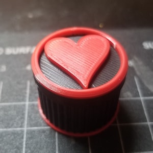 Peut inclure: Un bouchon de bouteille imprimé en 3D noir et rouge avec un motif en forme de cœur.
